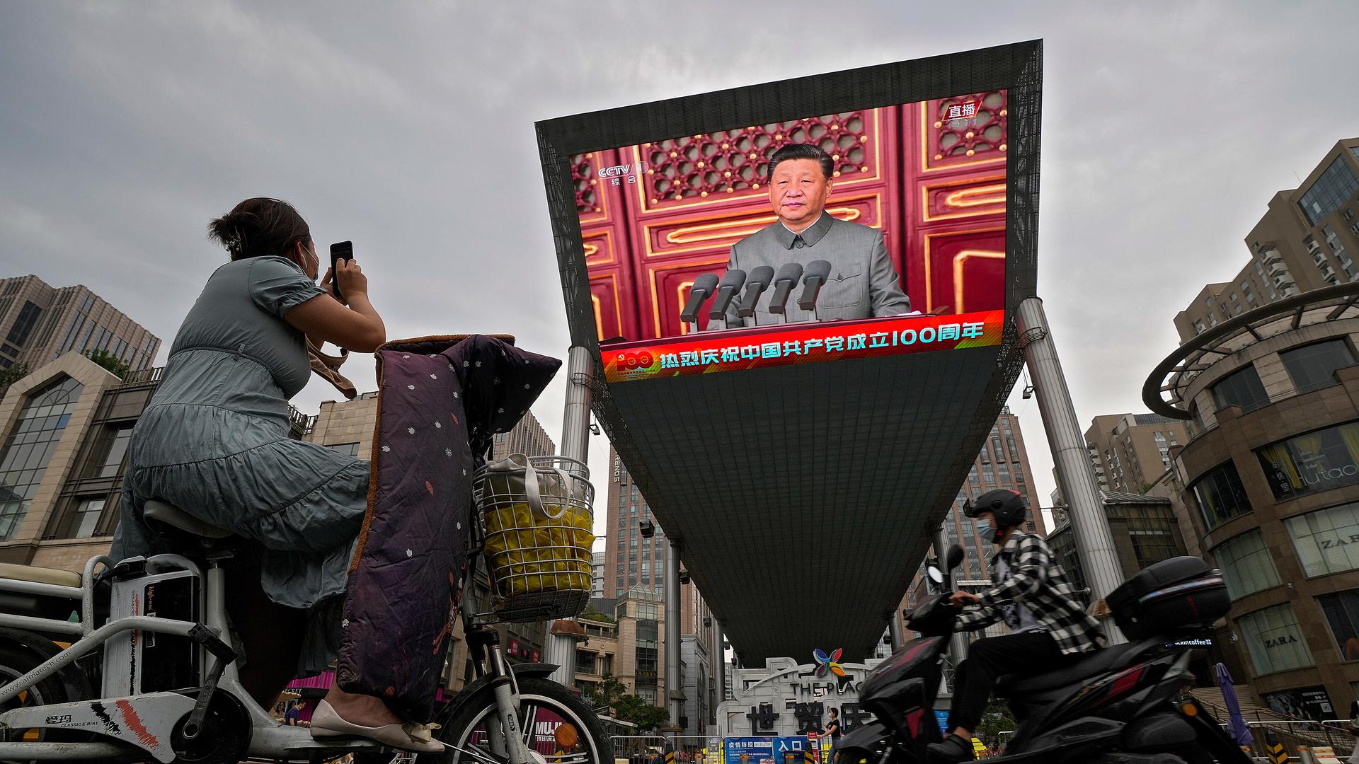 A woman on her electric-powered scooter films a large video screen outside a shopping mall showing Chinese President Xi Jinping speaking during an event to commemorate the 100th anniversary of China's Communist Party at Tiananmen Square in Beijing.