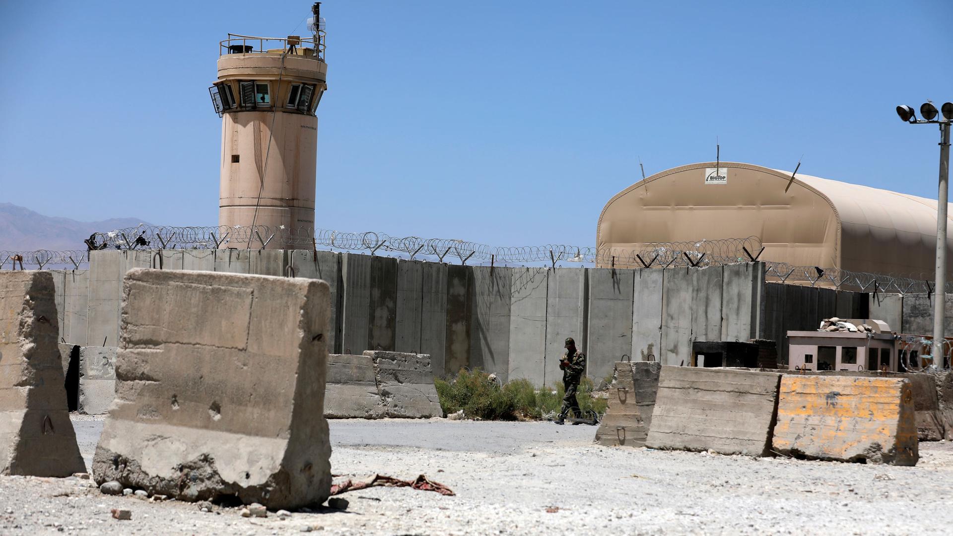A tall control tower is shown with large concrete barriers on the ground nearby with barbed wire on top.
