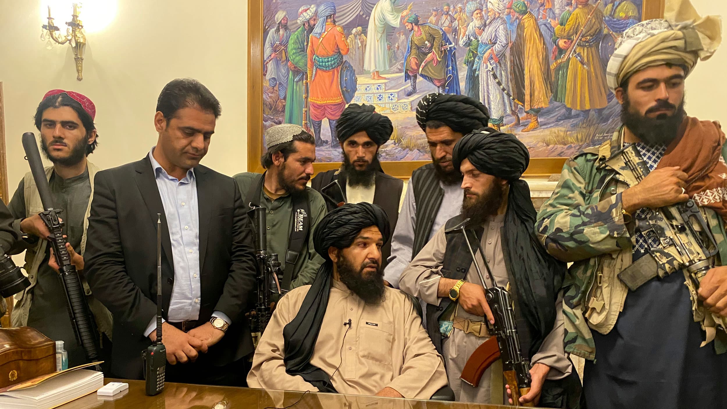 Several Taliban fighters are shown standing around a large wooden table with one man sitting down.