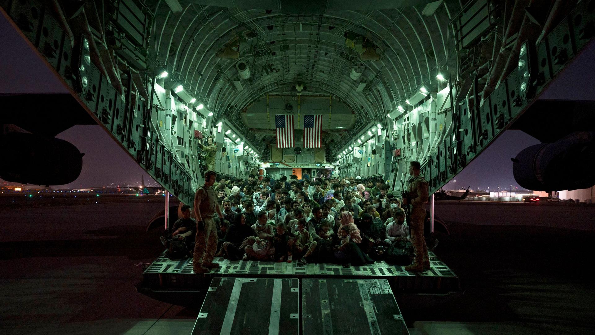 A large group of evacuees are shown in the back of a large Air Force aircraft with green-colored lights lining the inside.