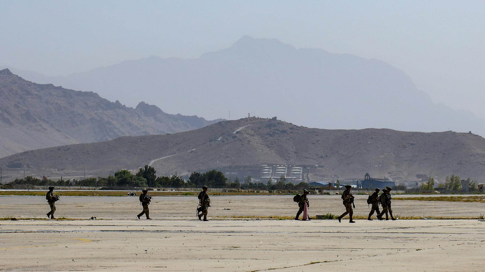 Several military officers are shown in a line, and heavily armed, walking on an open tarmac area with mountains in the distance.