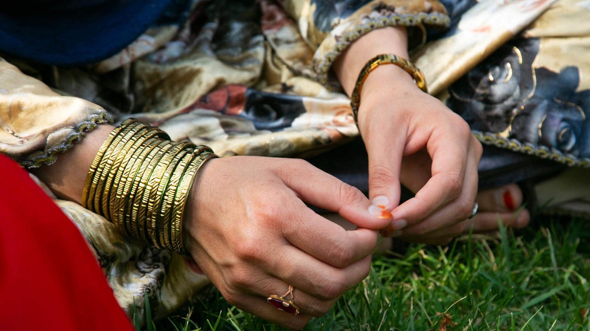A close-up photograph shows a woman's hands with lots of golden-colored braclets on her right wrist and a watch on the left wrist.