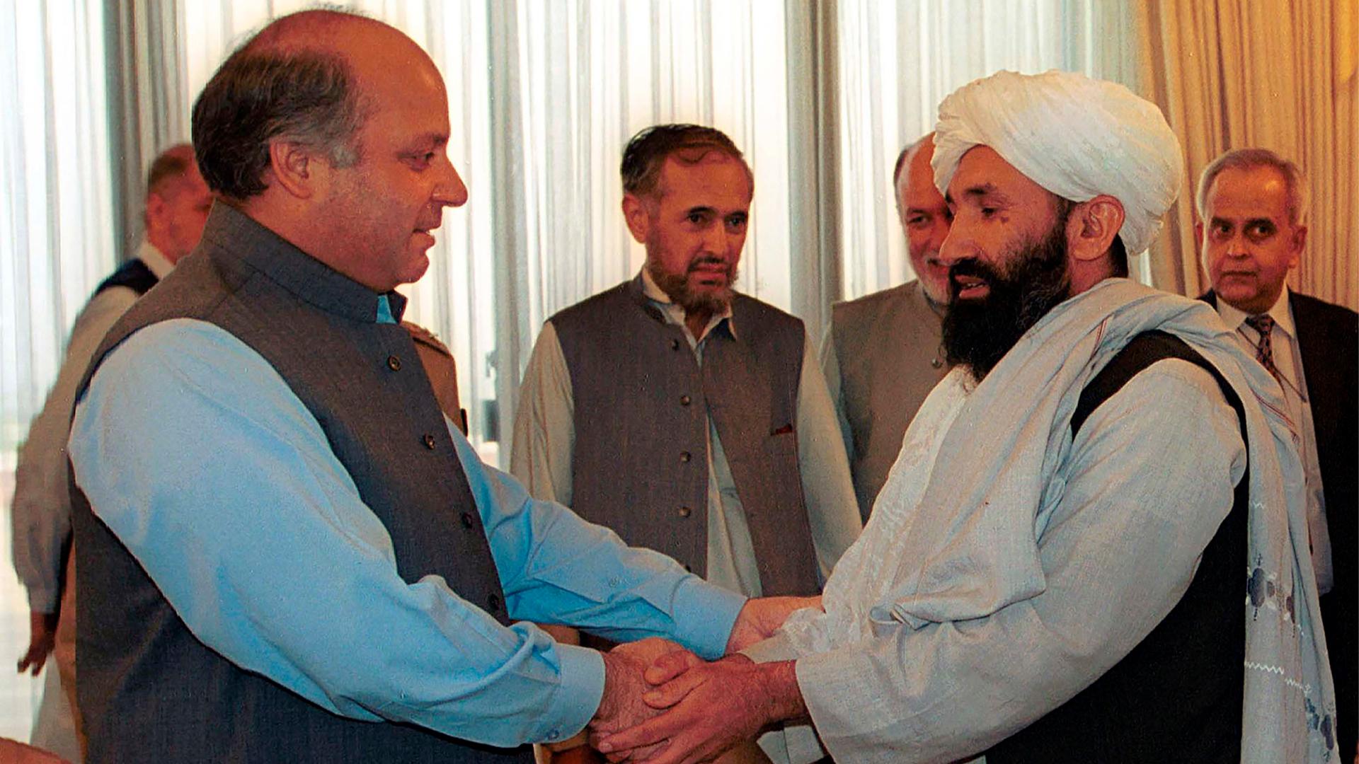 Mullah Hasan Akhund is shown wearing a white head wrap and standing across from Nawaz Sharif who is wearing a gray vest and blue shirt.