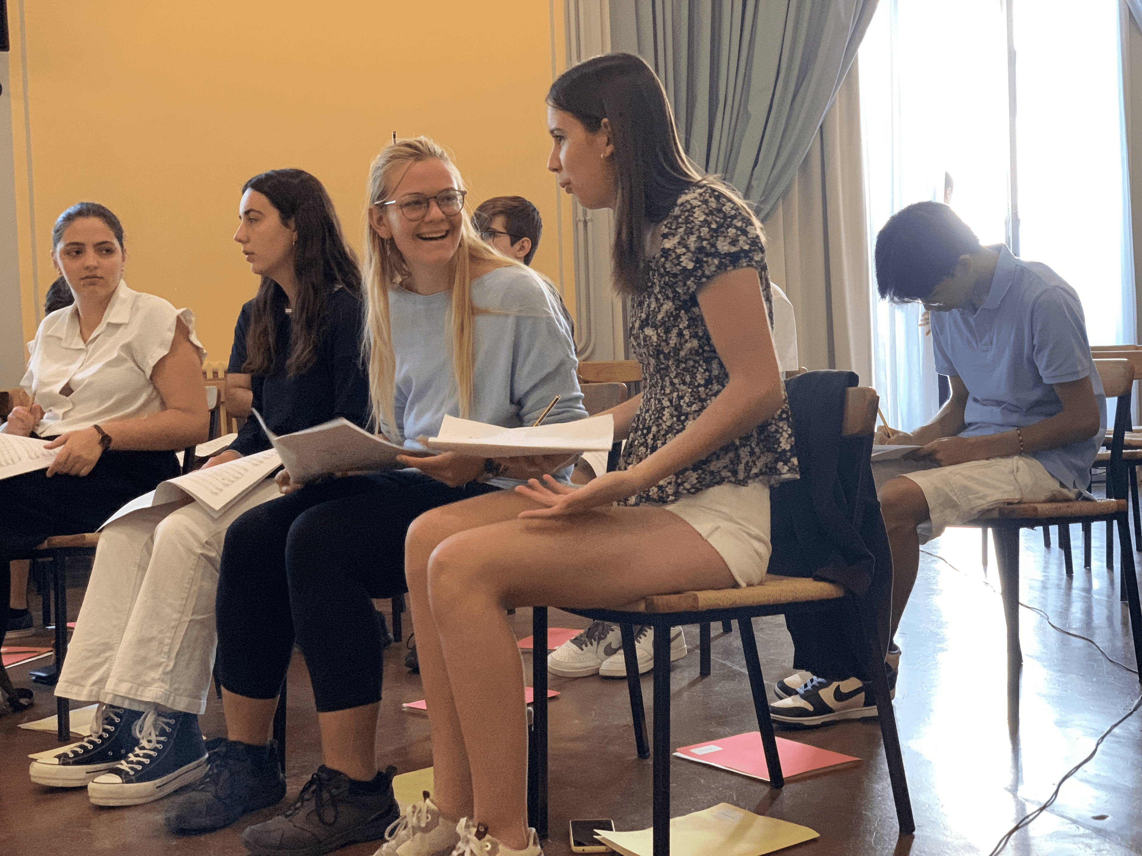 Various young women and men sitting in a room and holding papers to rehearse