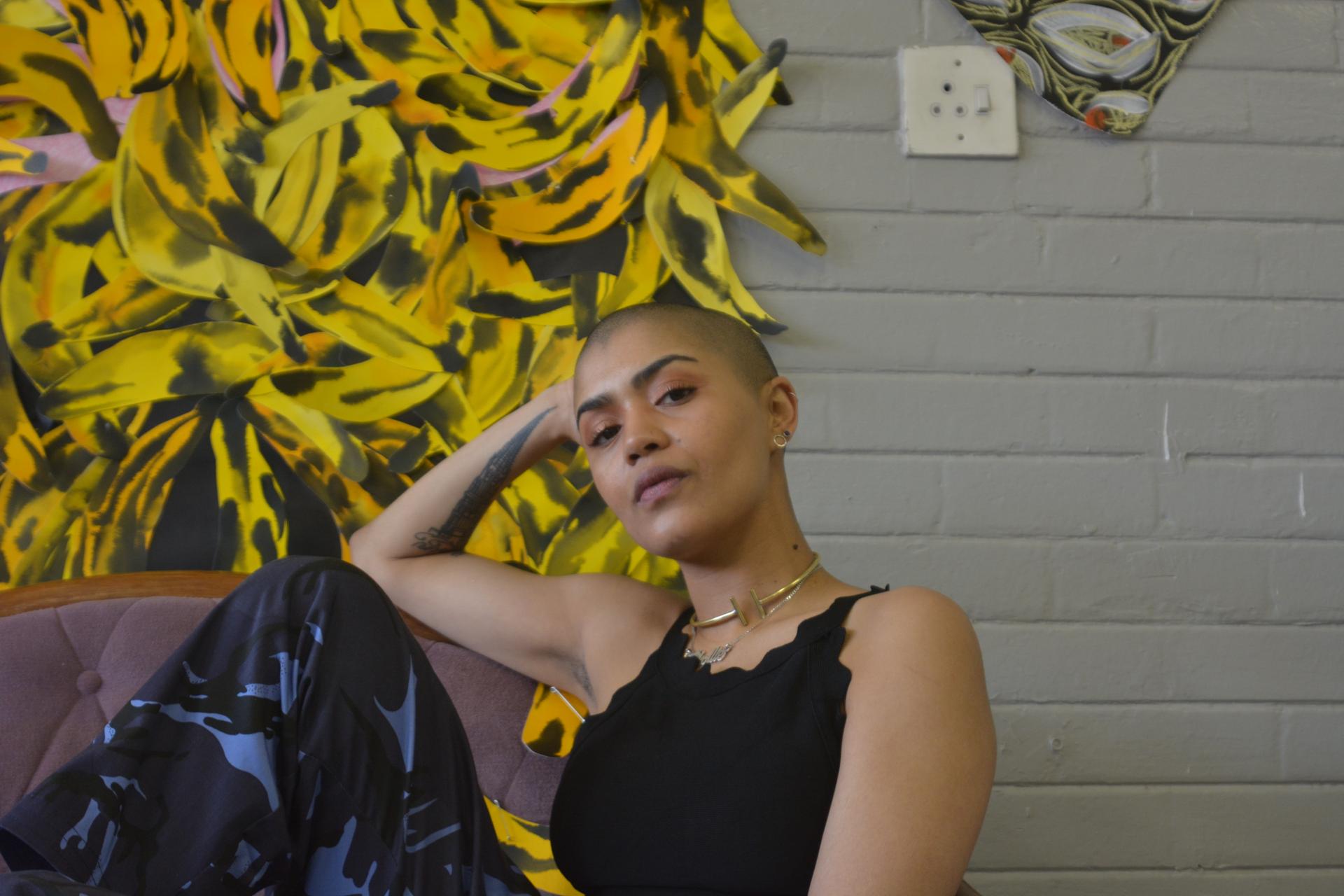 South African artist Lady Skollie on a sofa in her studio with cut-out shapes of bananas on the wall behind her