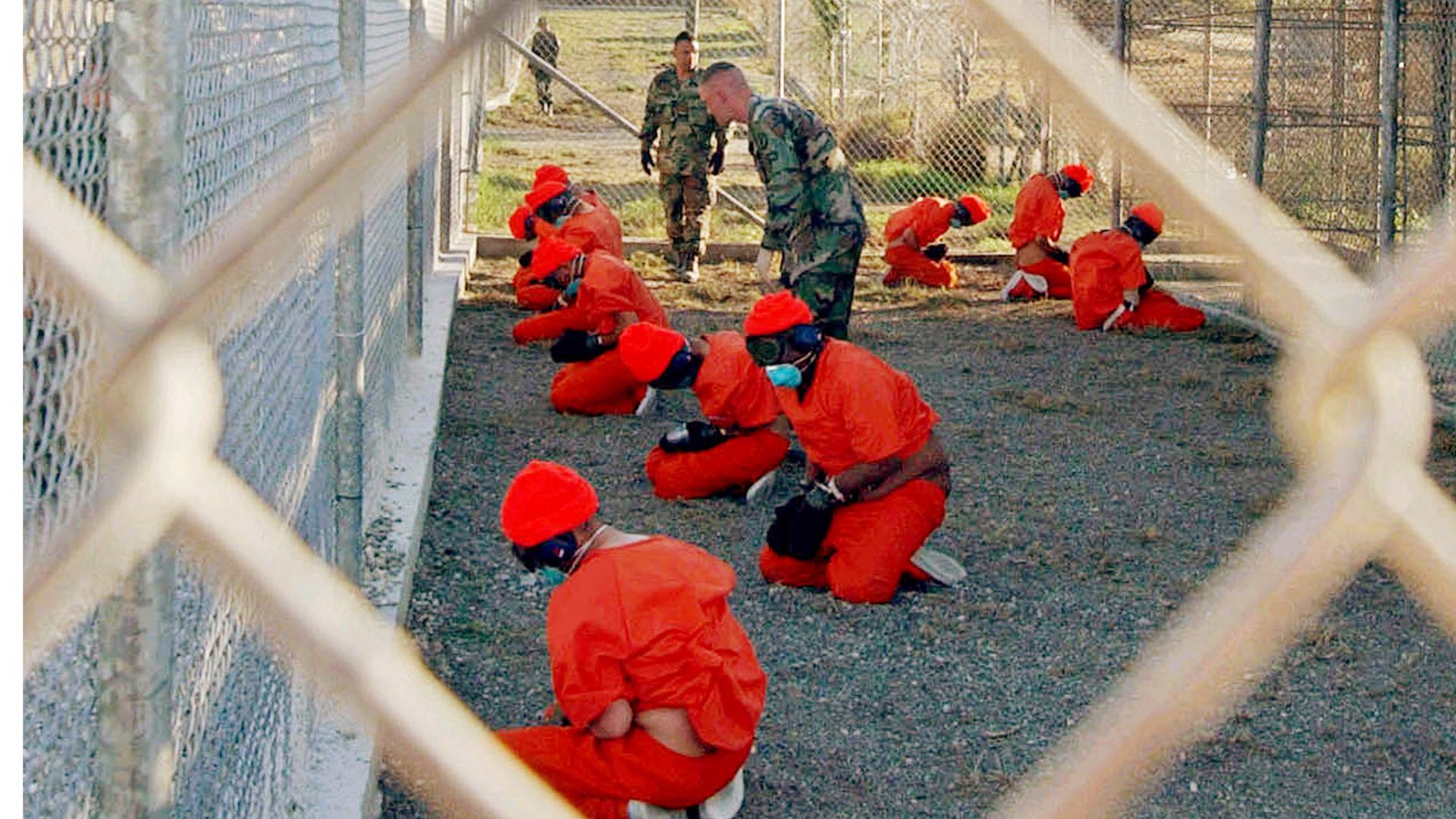 Detainees in orange jumpsuits sit in a holding area watched by military police at Guantanamo Bay's Camp X-Ray in 2002.