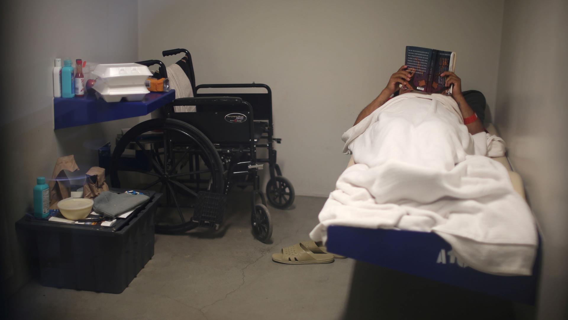 Man lies in bed in small room with wheelchair next to him