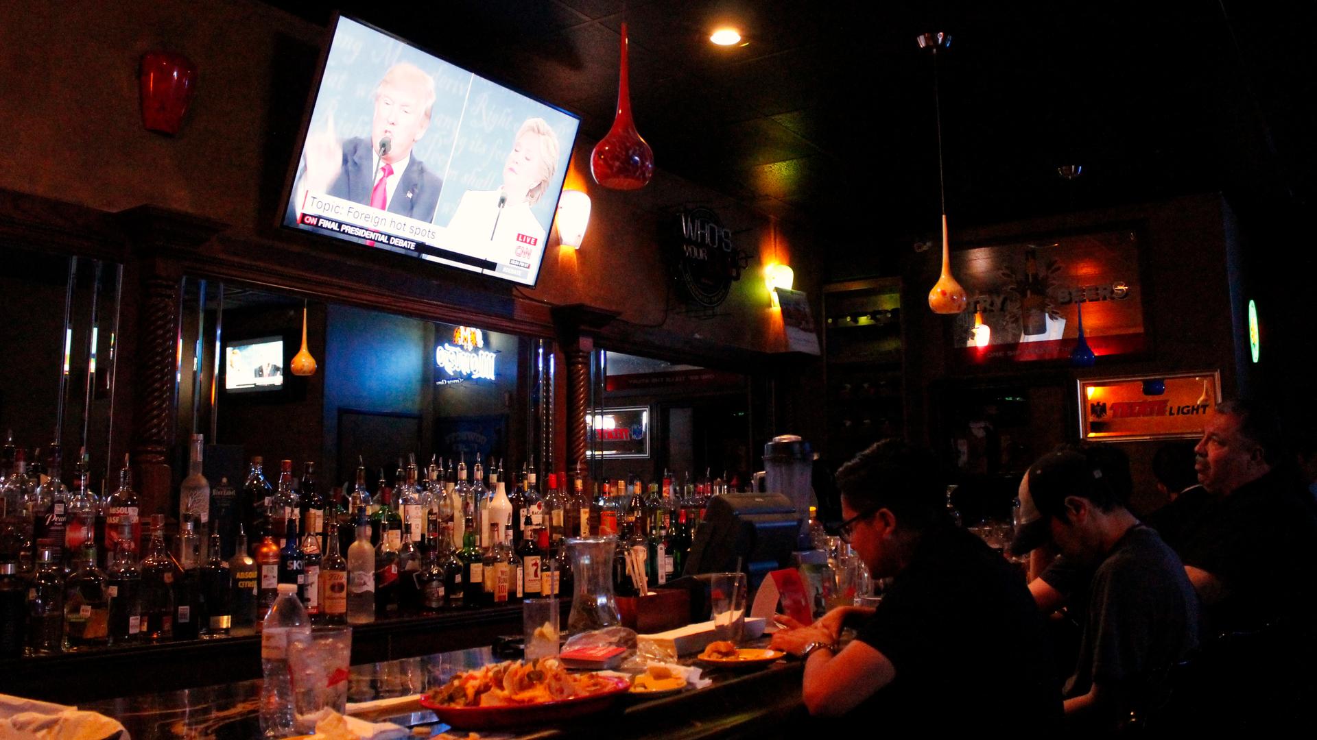 People sitting at a bar with a screen showing Donald Trump and Hillary Clinton above