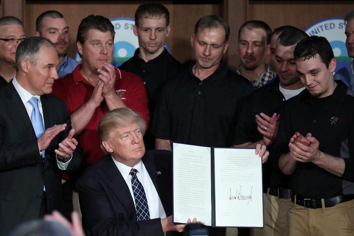 A group of miners wearing black polos and several onlookers watch President Donald Trump as he holds up a signed version of his most recent executive order, “Promoting Energy Independence and Economic Growth.”