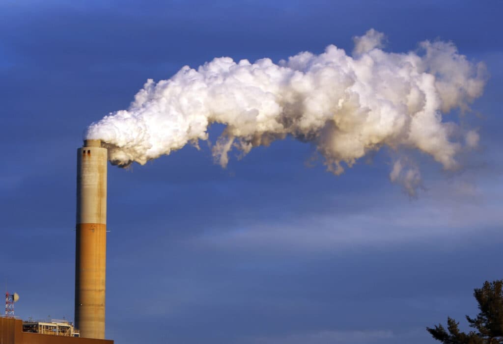 A tall smokestack emitting a large plume of white smoke against a clear blue sky.