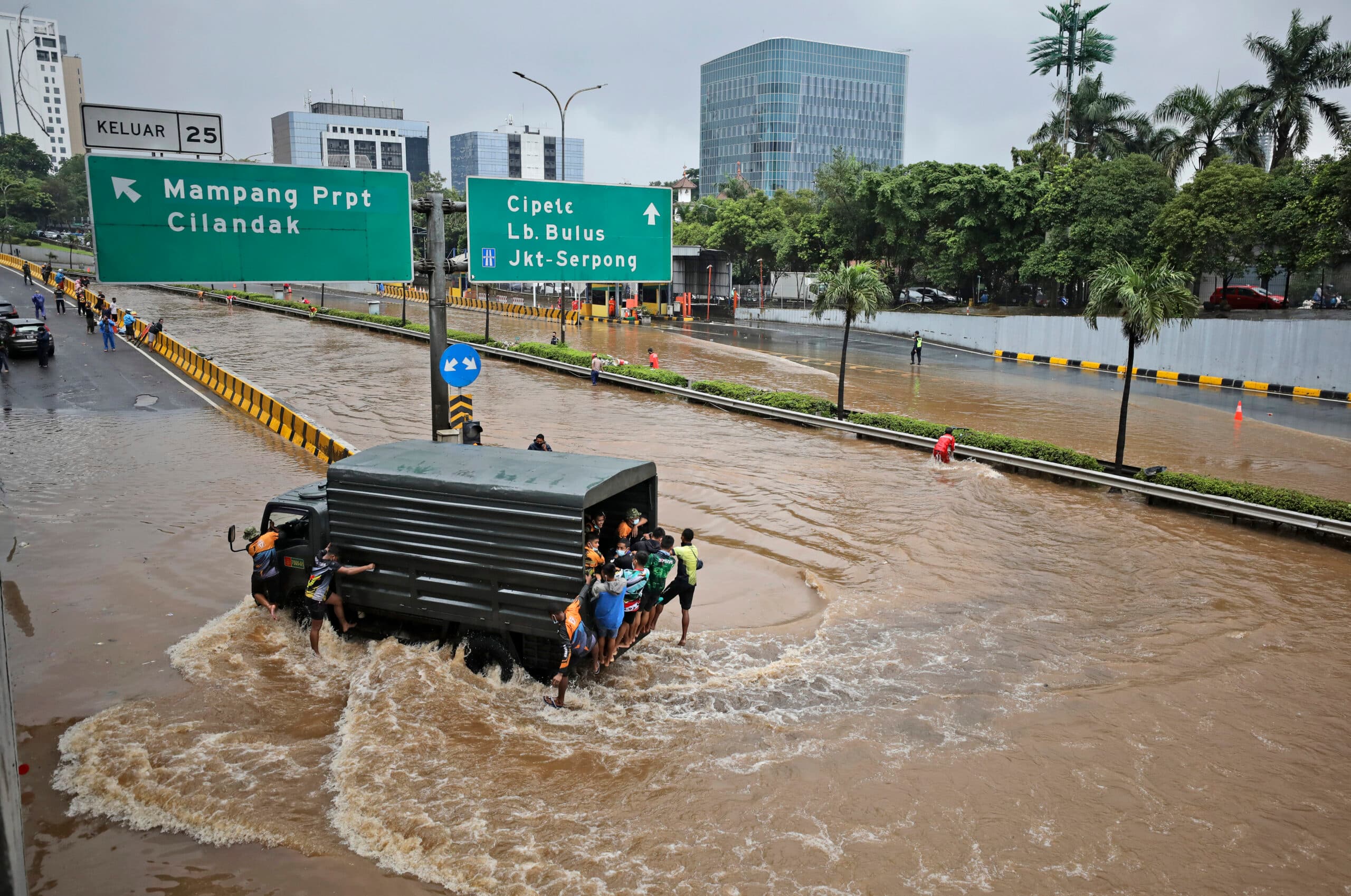 A flooded city road with a military truck carrying people through the water, while others wade alongside. Overhead signs indicate directions to various city areas. The surrounding area has buildings and trees with a cloudy sky.