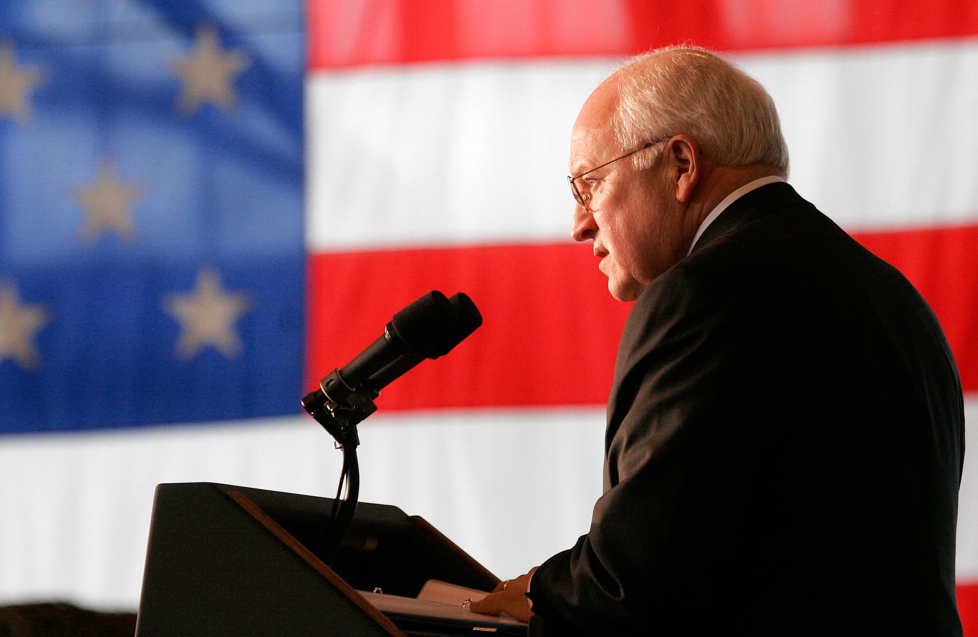 A man in a suit speaking at a podium with an American flag backdrop.