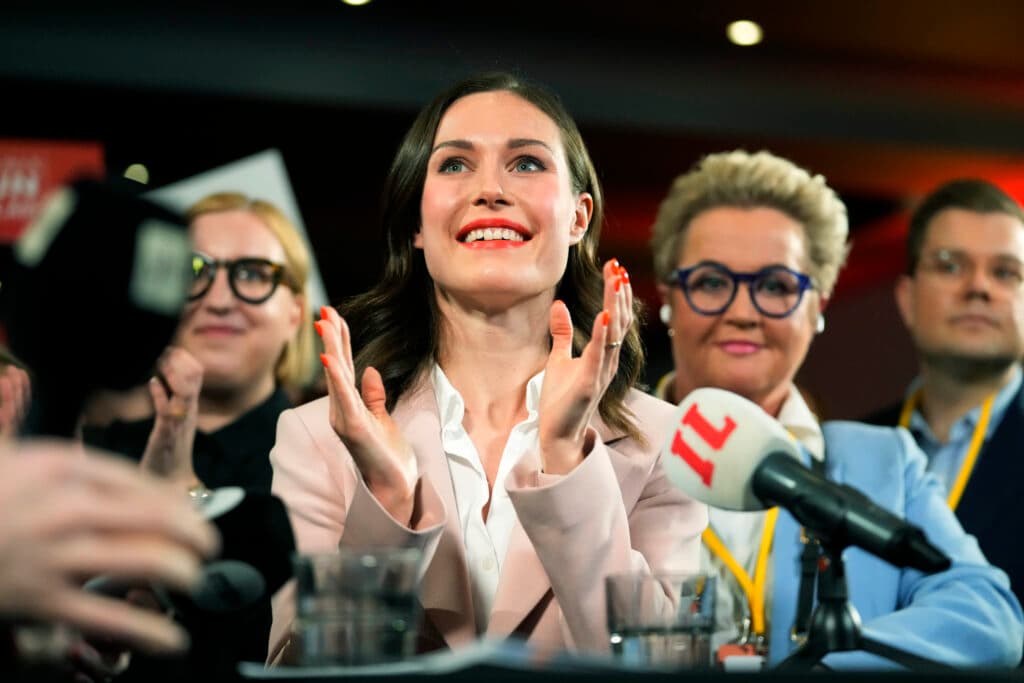 A woman in a pink blazer claps her hands enthusiastically in front of microphones, surrounded by several people in a well-lit event setting.
