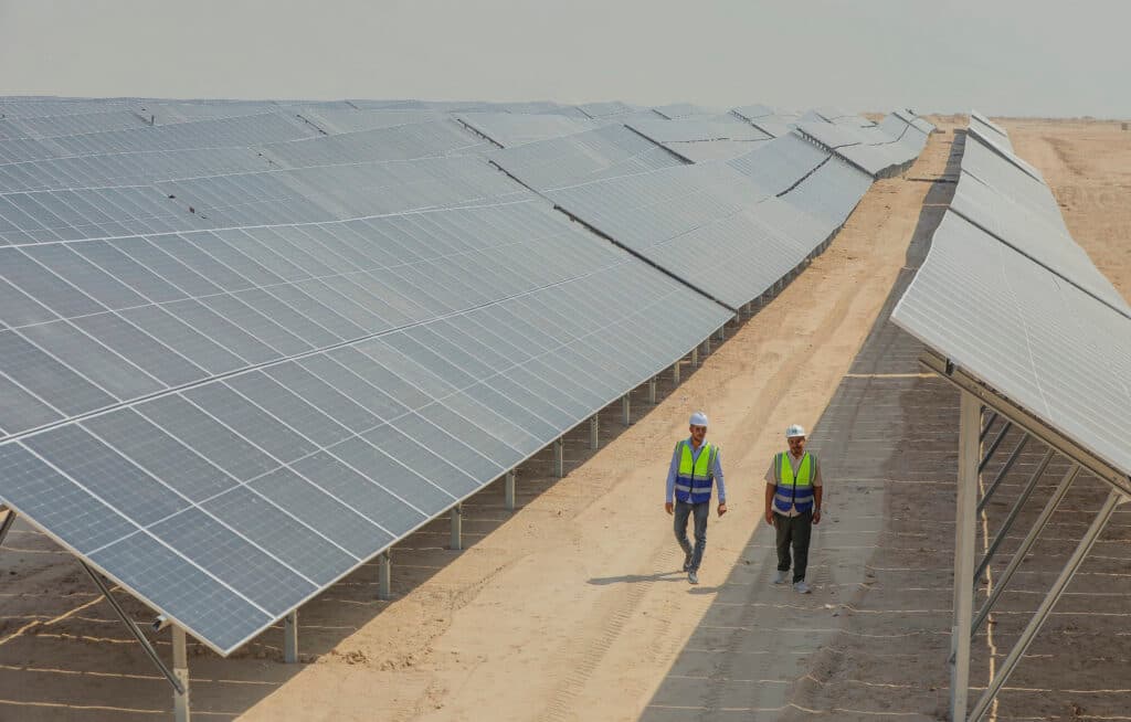 Two workers in safety vests and helmets walk between rows of solar panels in a desert-based solar farm.