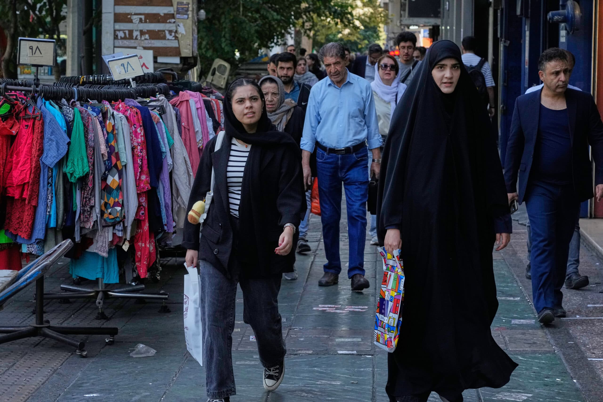A group of people walking along a city street next to a clothing stall displaying colorful garments. The scene includes women wearing different styles of head coverings, and a variety of street fashion. Trees and shopfronts are visible in the background.