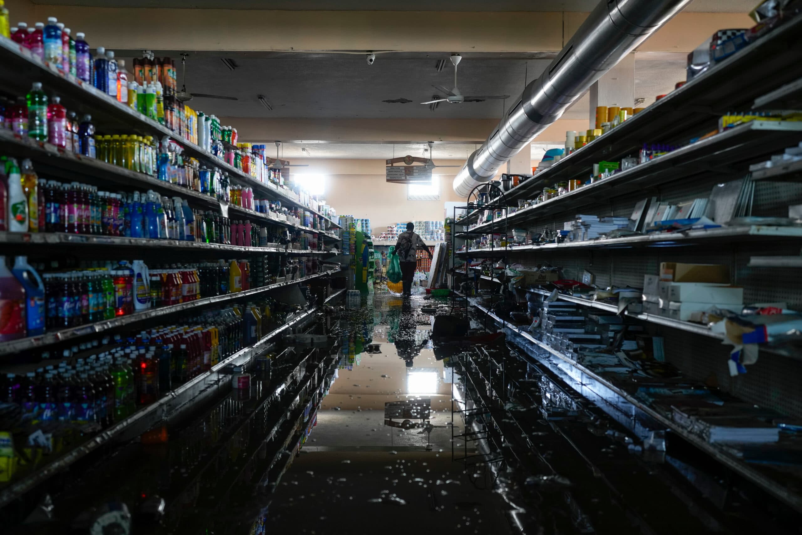 A dimly lit supermarket aisle with shelves full of products is flooded with water. A person is seen in the distance holding a bag, walking carefully through the water.