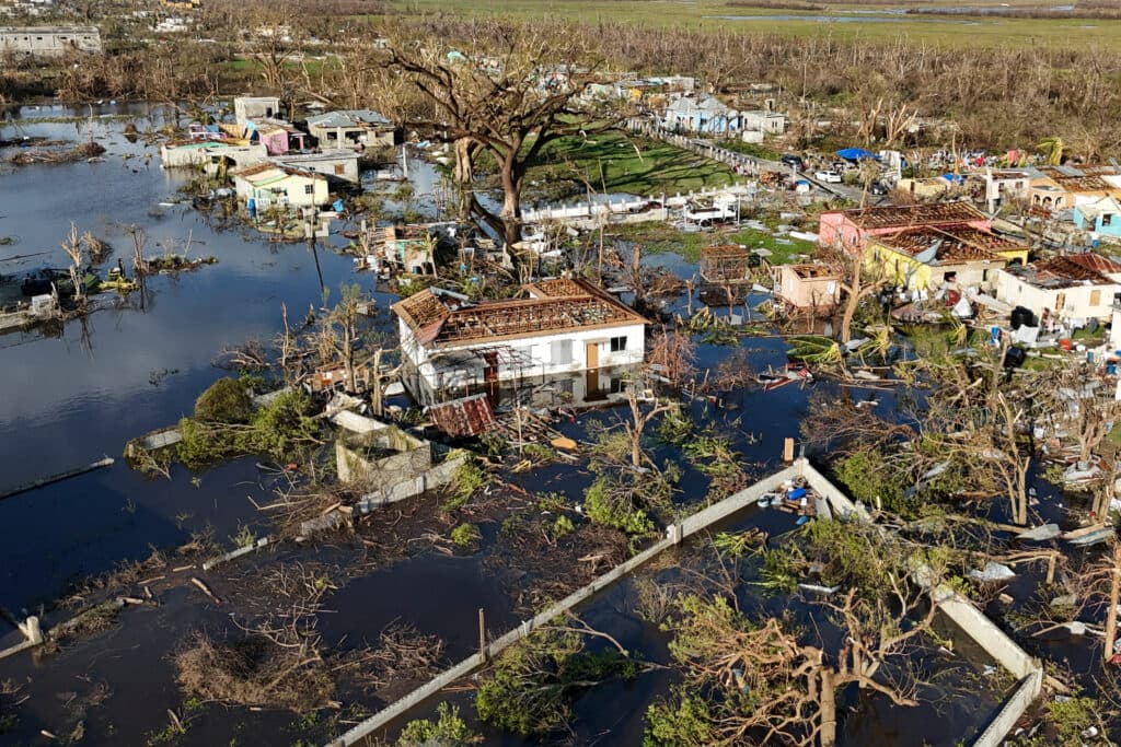 Aerial view of a flood-ravaged area with partially submerged houses, damaged roofs, and fallen trees, surrounded by murky water and debris.