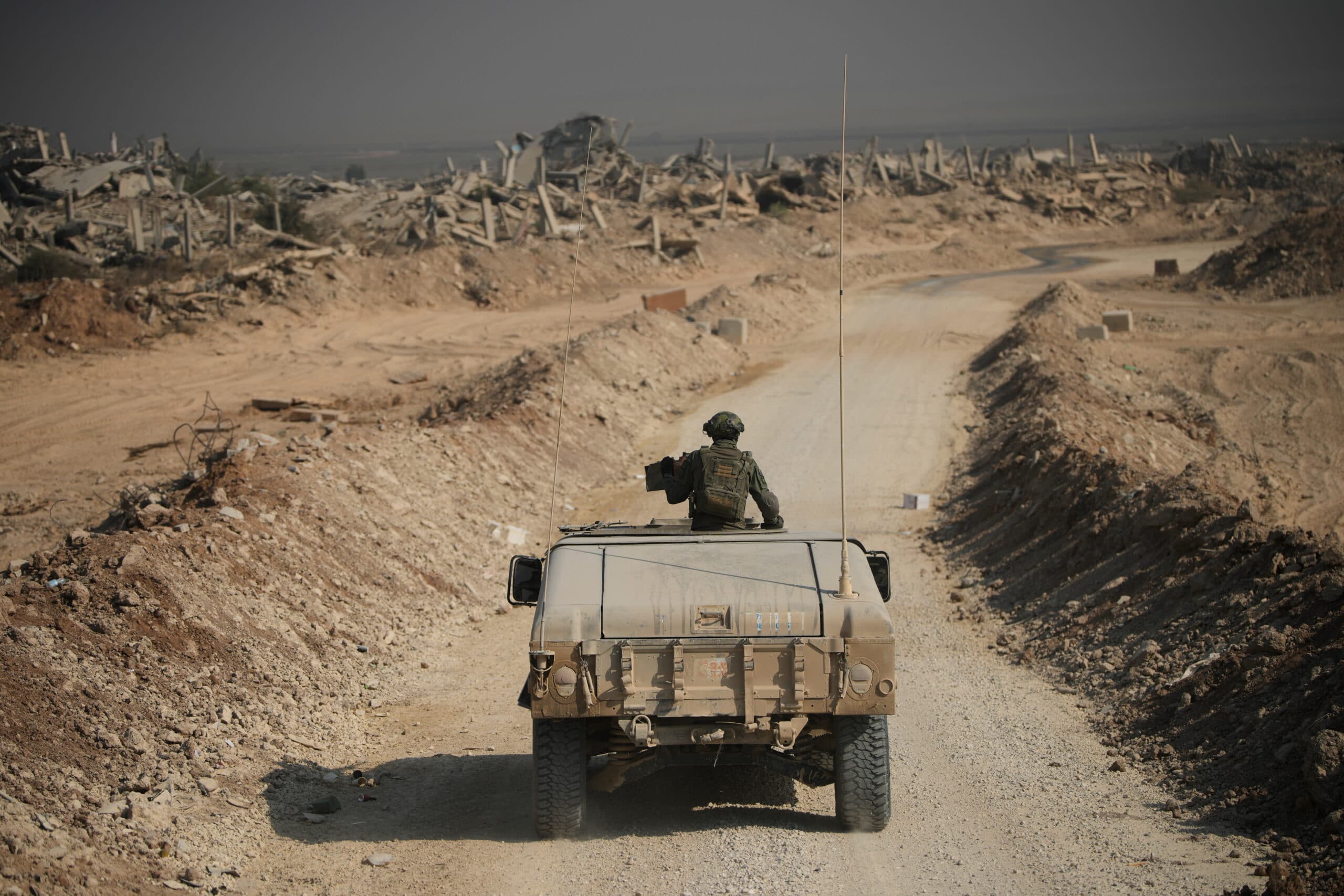 A soldier in camouflage sits atop an armored vehicle driving along a dirt road surrounded by a desolate landscape, with the remains of destroyed buildings in the background.