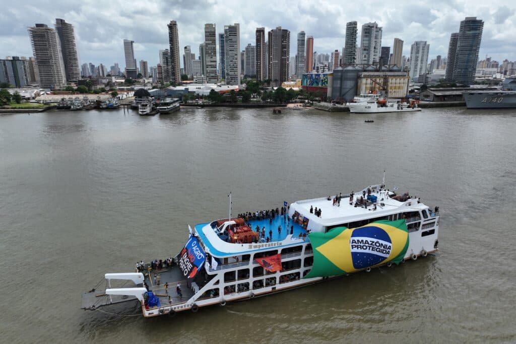 A riverboat with a large Brazilian flag design and the text "Amazônia Protegida" on its side, navigating a river in an urban area. The boat is carrying numerous people and is decorated with banners. A skyline of tall buildings and other vessels is visible in the background under a cloudy sky.