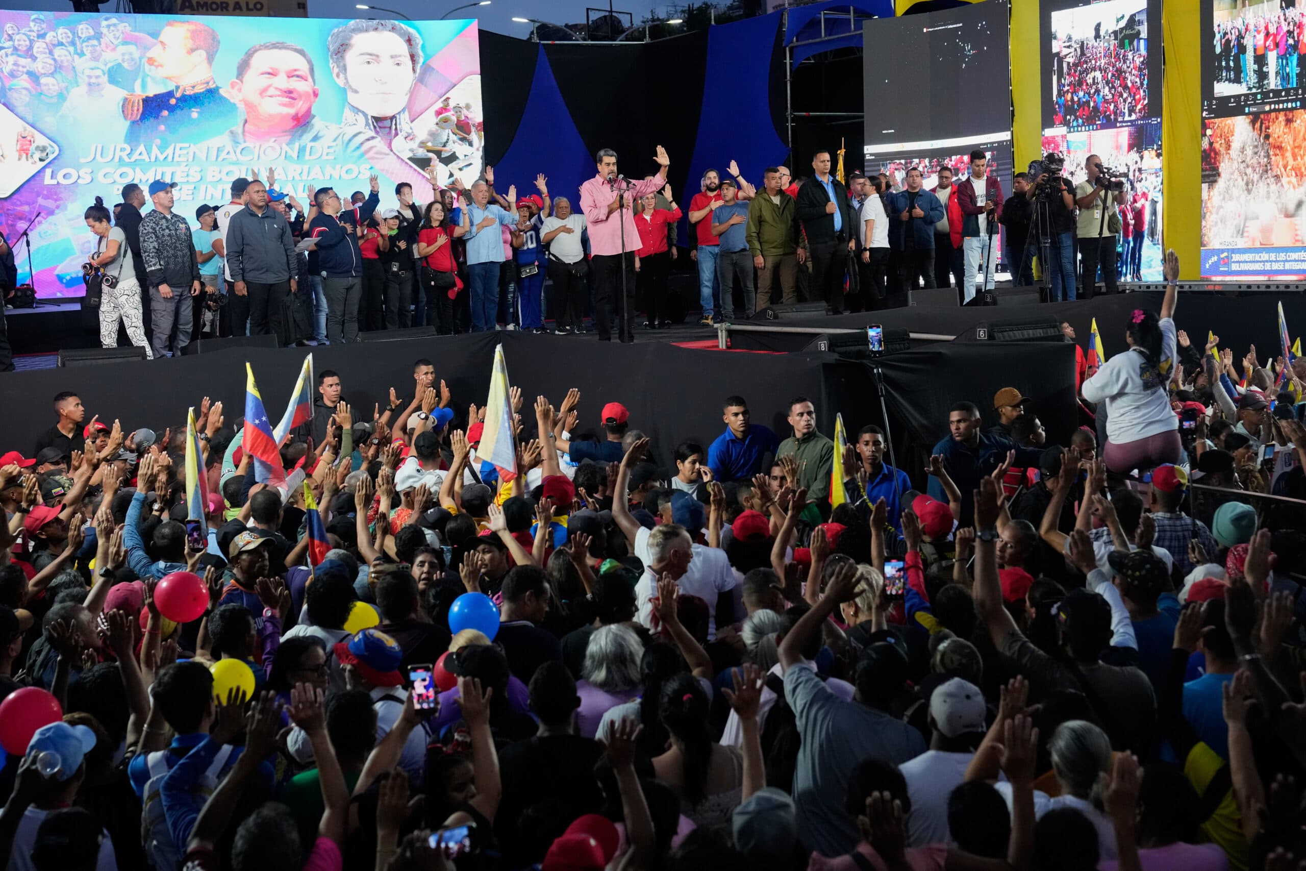 A large crowd gathered at an outdoor event with many people raising their hands. A speaker stands on stage with a group of people behind a podium. The backdrop includes large images and text in Spanish, surrounded by video screens and colorful decorations.