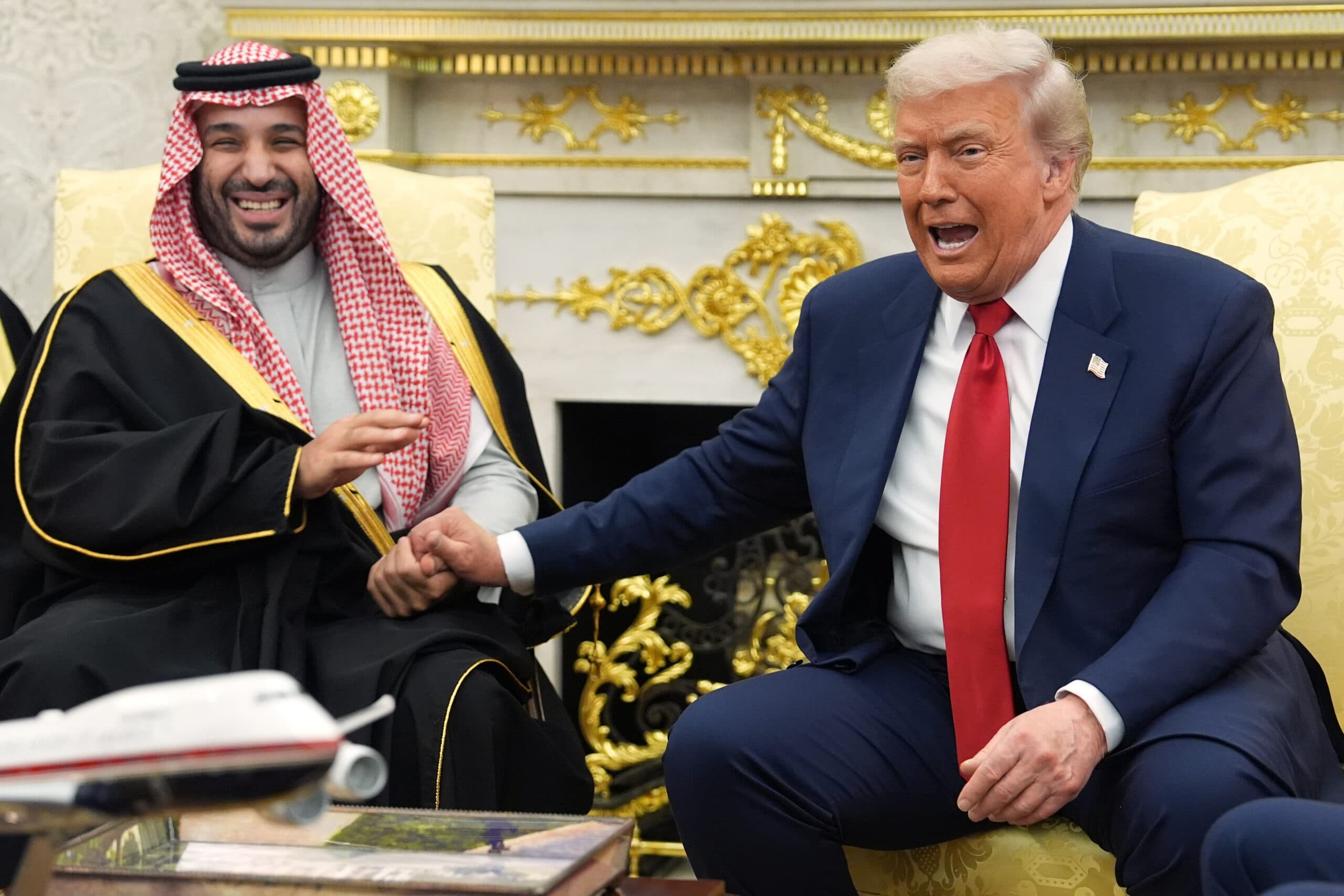 Two men in formal attire sitting in a lavish, gold-decorated room, smiling and sharing a friendly gesture, with a model airplane on the table in the foreground.