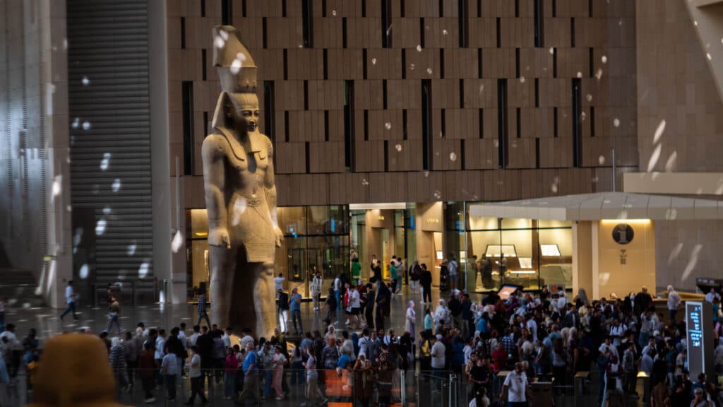 A large indoor area with a towering ancient Egyptian statue surrounded by a crowd of people. The statue depicts a standing figure with traditional headdress and attire. The area features a high ceiling with patterns on the walls, and the lighting casts reflections across the space.