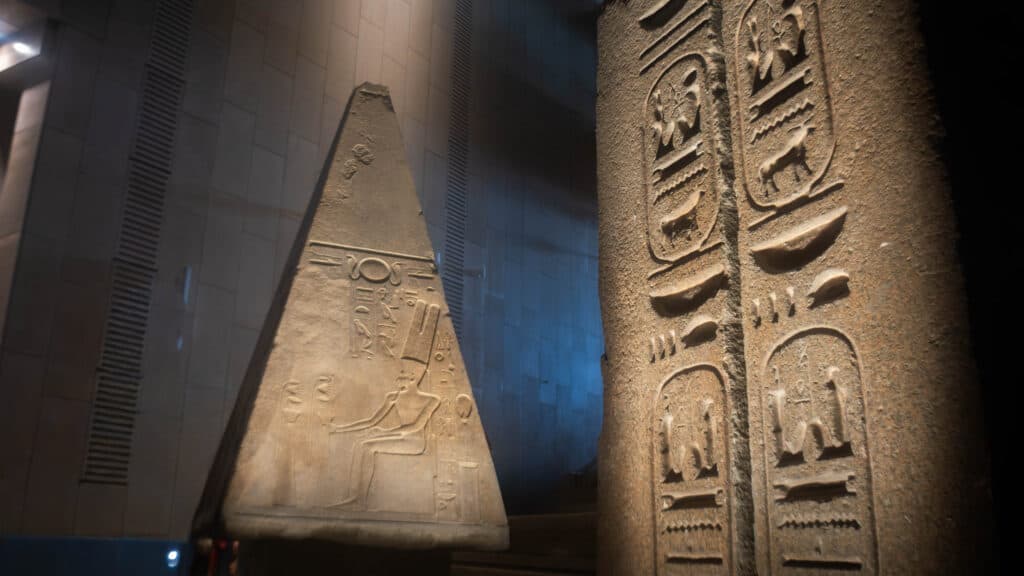An ancient Egyptian stone artifact featuring hieroglyphics, with a pyramid-like shape in the background and intricate carvings on a vertical surface in the foreground, displayed in a dimly lit museum setting.