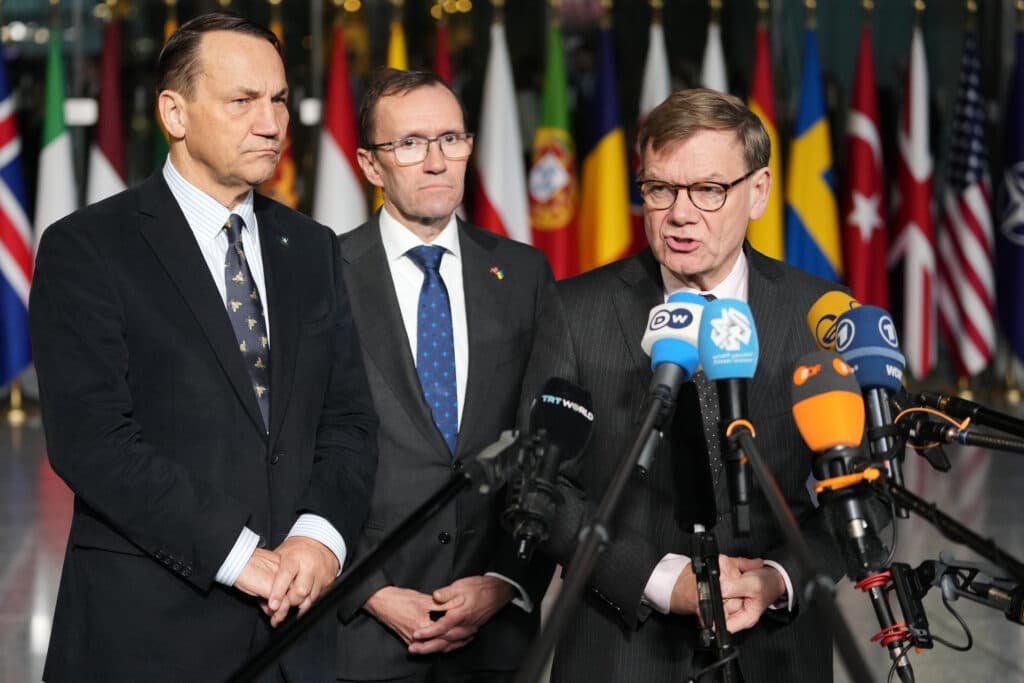 Three men stand in front of microphones during a press conference, with various country flags visible in the background.