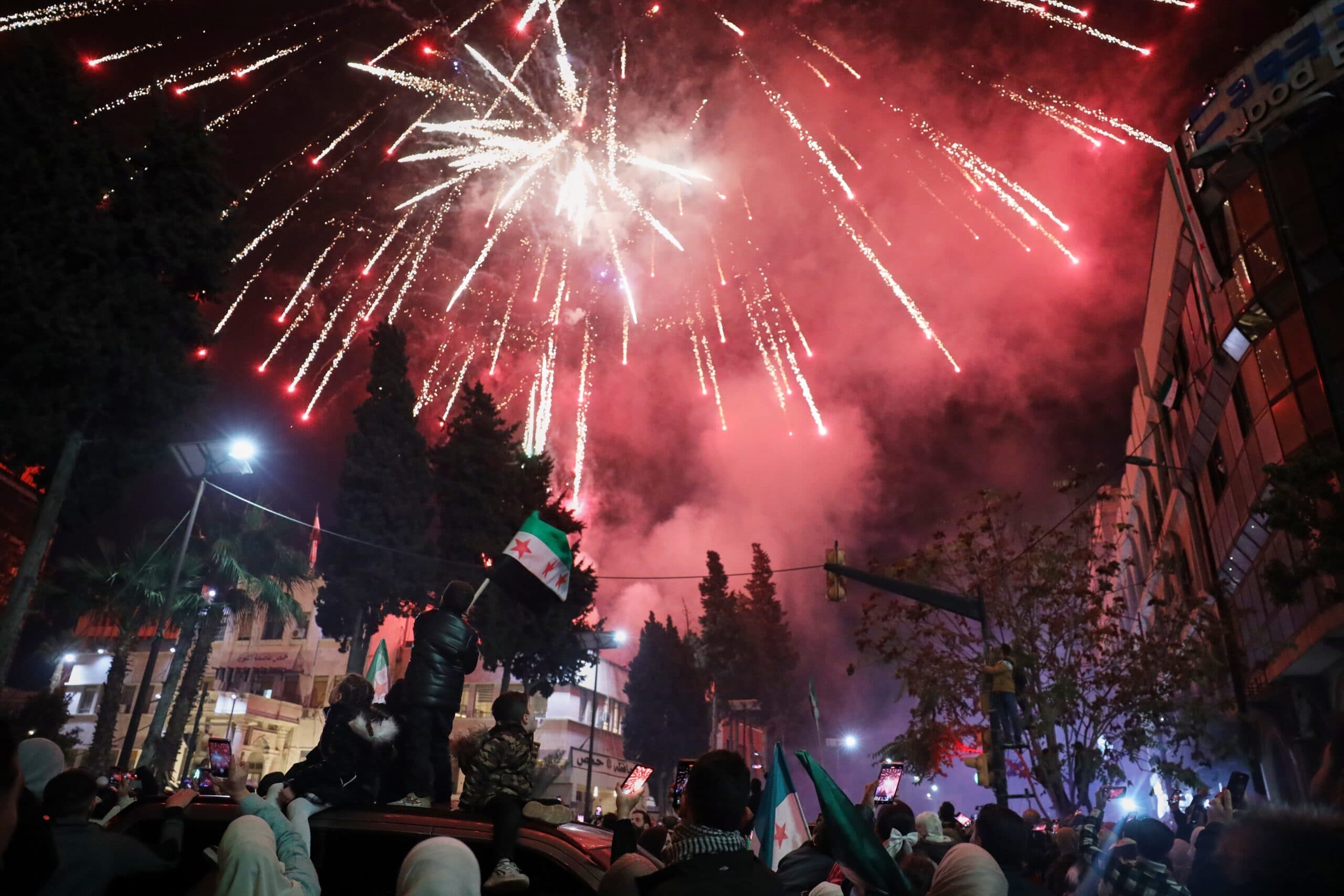 A crowd of people celebrating with fireworks lighting up the night sky, while some individuals wave flags and take photos.