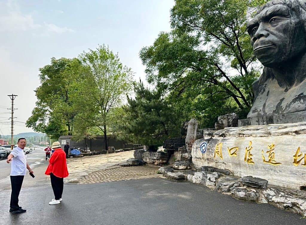 Two people walking on a path next to a large sculpture of a hominid head in a park, with trees and a stone wall bearing Chinese characters.
