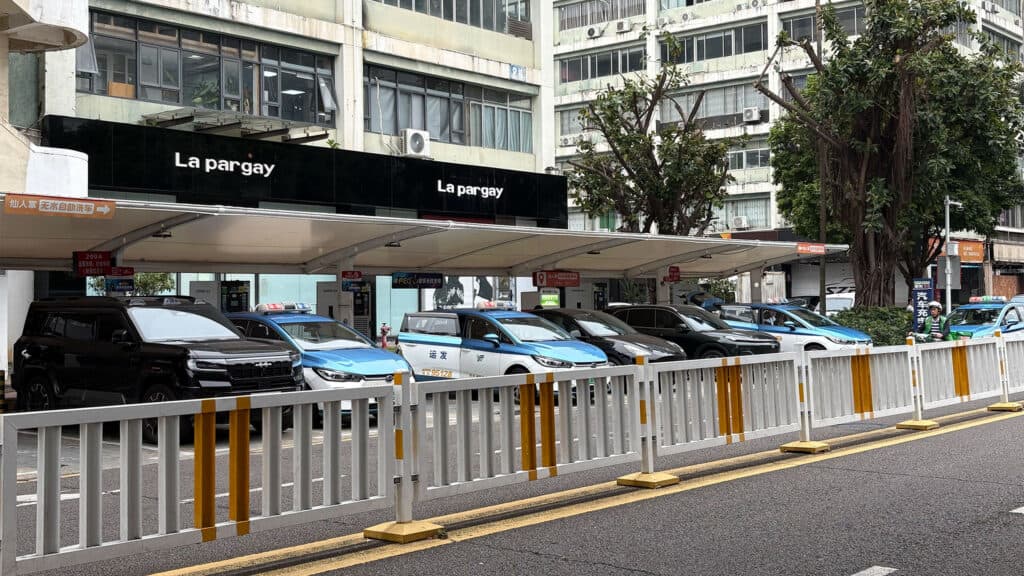 A row of parked vehicles under a covered car park with a black sign above reading 'La pargay'. The building behind has multiple windows and there are trees visible to the right. A white and yellow traffic barrier runs along the front.