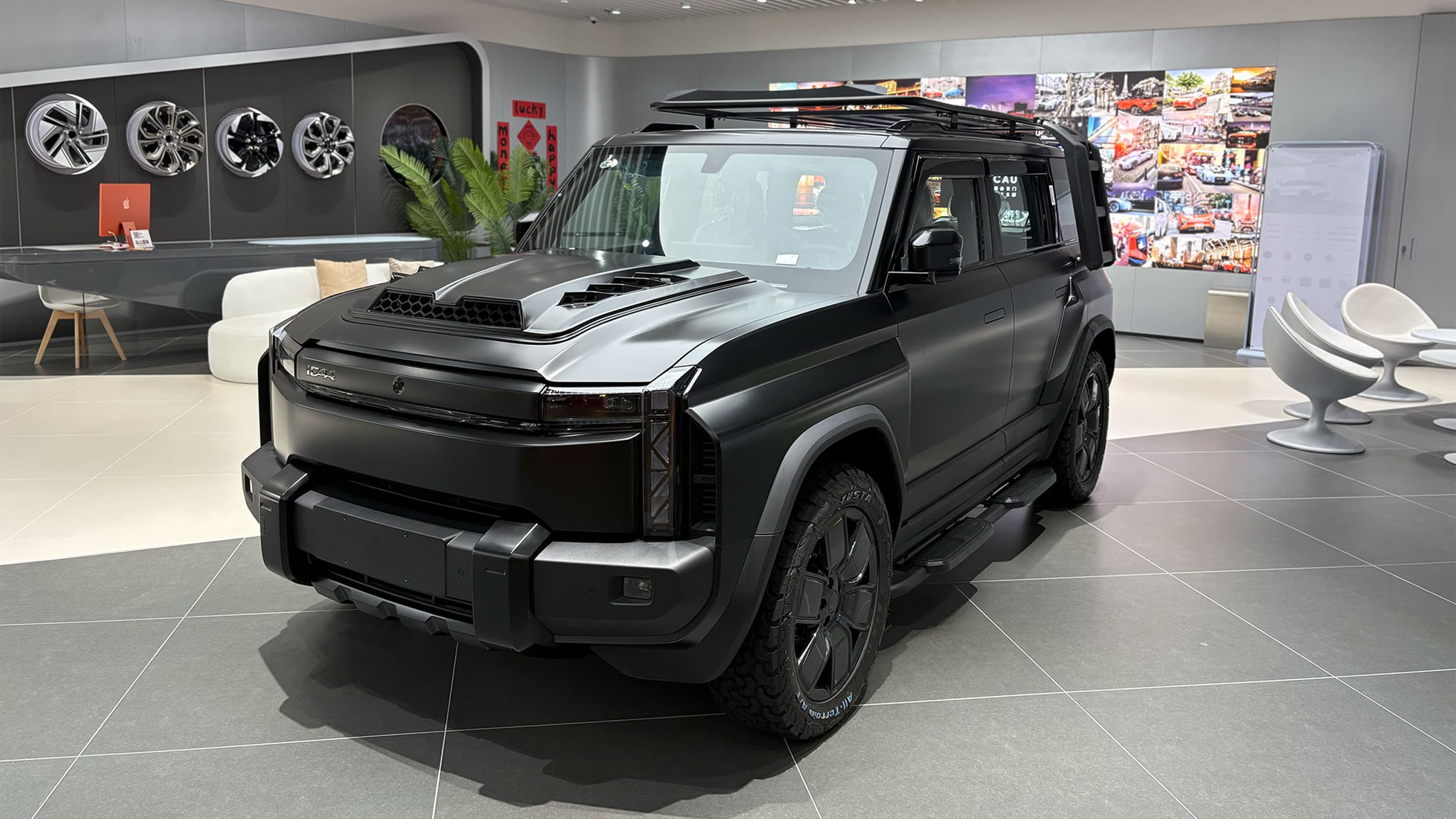 A sleek black SUV with a rugged design on display in a modern showroom, featuring prominent wheel arches, a distinctive hood with air vents, and a roof rack. The room has a tiled floor, decorative elements, and wheels mounted on the wall in the background.
