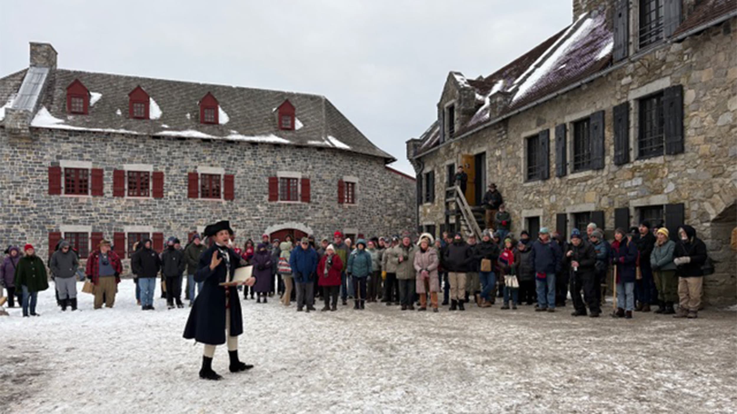 A person in historical clothing stands in front of a group of people gathered outside two stone buildings on a snowy day.