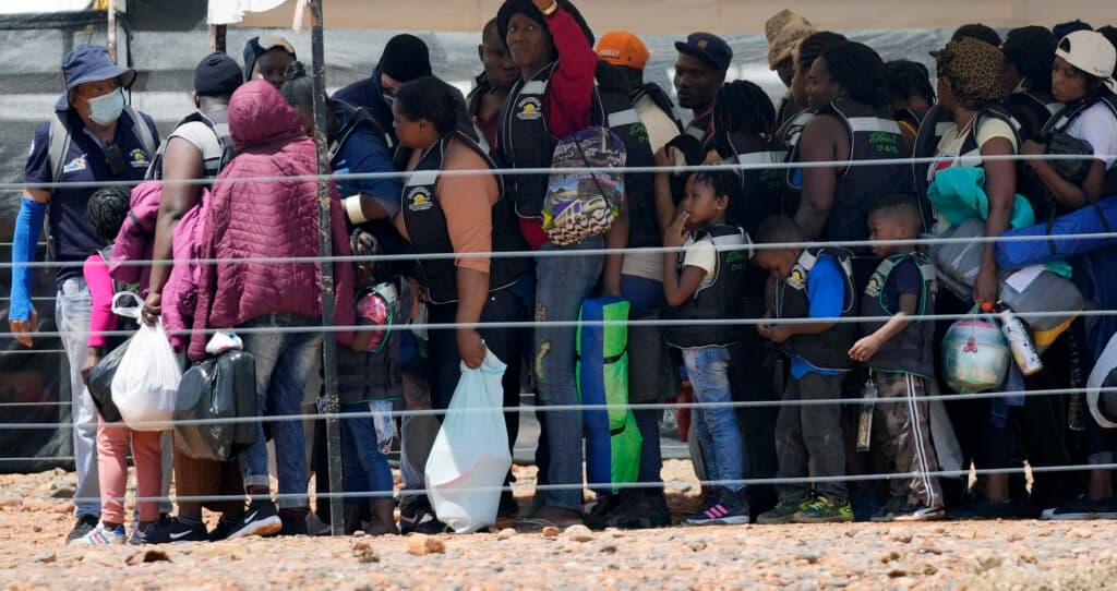 A group of people, including adults and children, are gathered closely together wearing life jackets. They are carrying various bags and appear to be in an outdoor setting with rocky ground and some fencing visible.