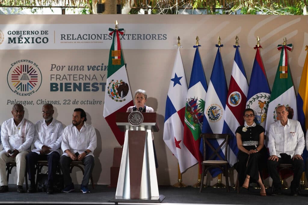 A speaker at a podium with the Mexican flag speaks at an international event in front of a sign that reads "Encuentro de Palenque" and several national flags, including those from Mexico, Panama, El Salvador, Costa Rica, Belize, and others. Seated individuals are dressed in formal white clothing, and the event appears to be a diplomatic meeting in Palenque, Chiapas, on October 22.