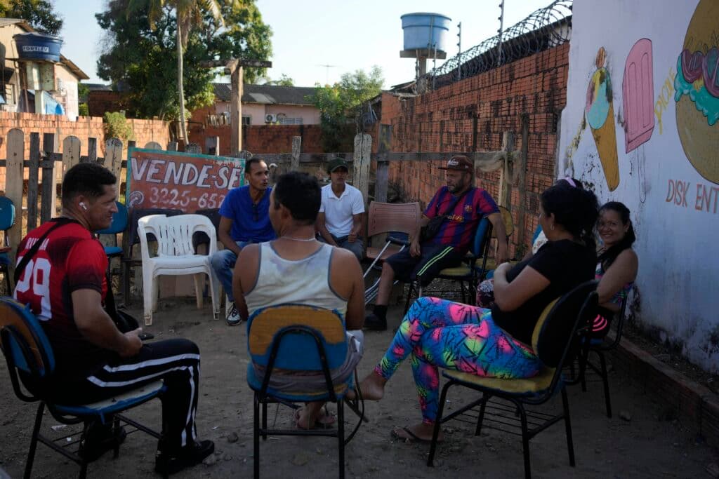 A group of people sitting in a circle outdoors, surrounded by a fence and brick walls. Some are wearing casual clothing, including a man in a sports jersey and a woman in colorful leggings. There's a sign in the background that reads 'VENDE-SE' next to a painted wall with drawings of food items.