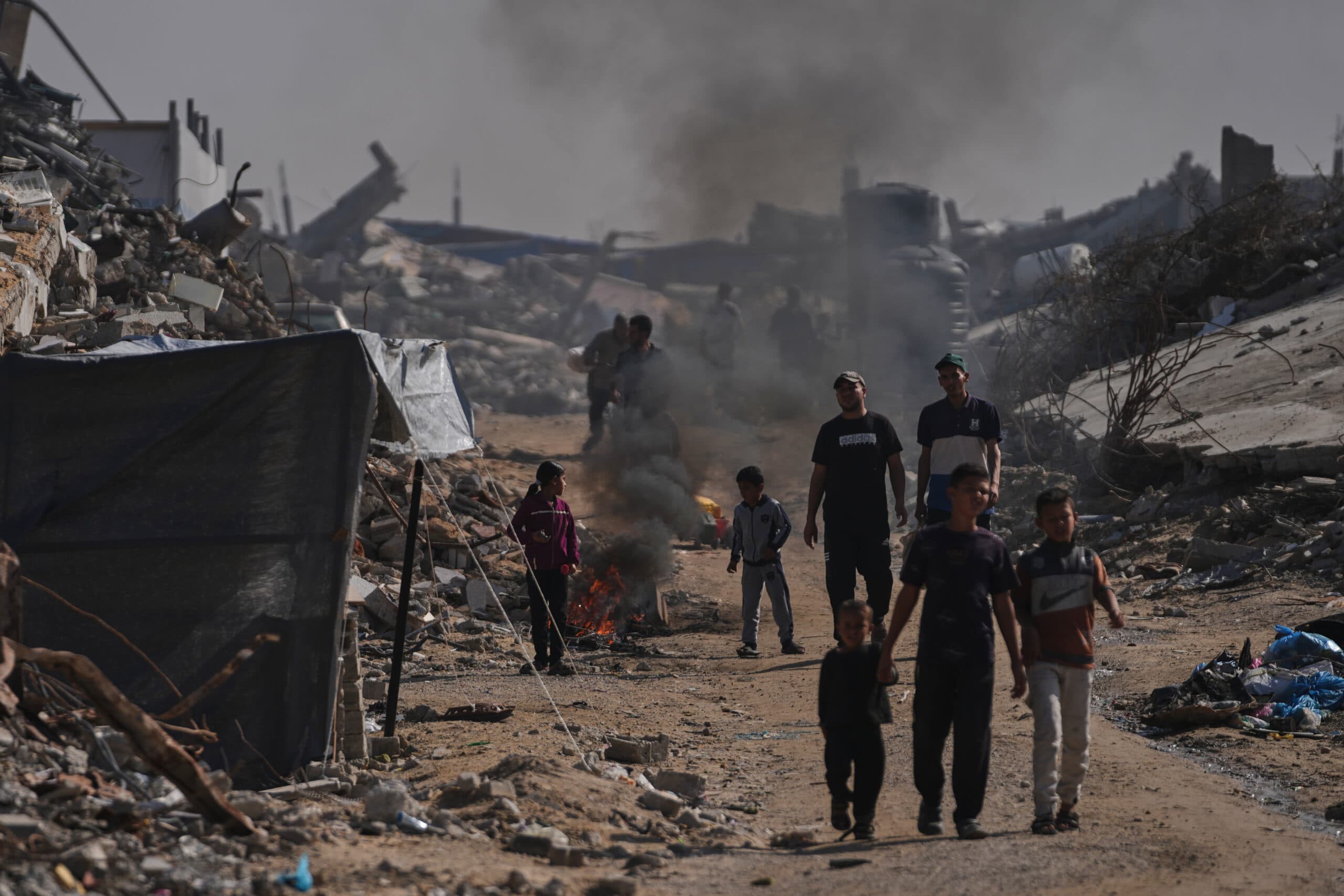 A group of people, including children, walk through a street littered with debris and rubble, with smoke rising in the background.