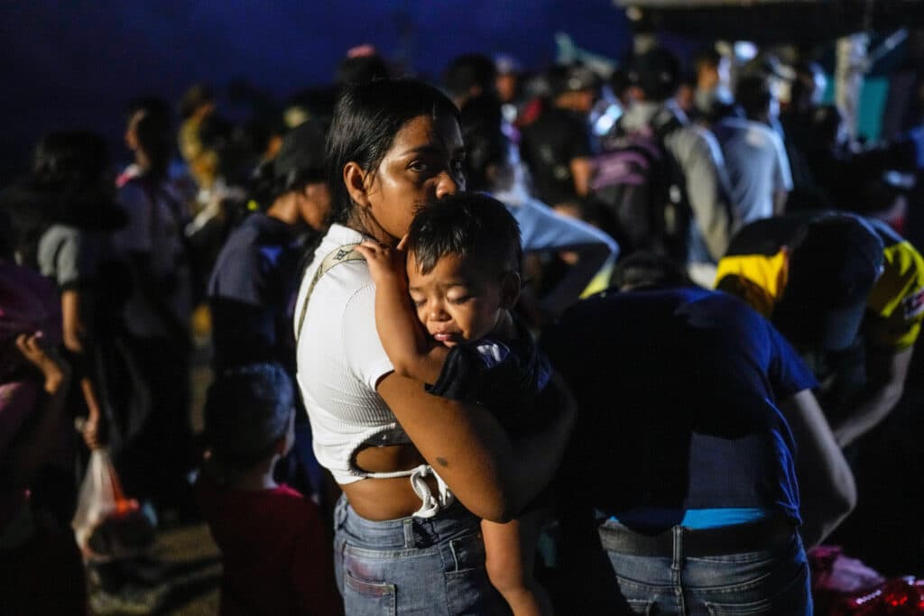 A woman holding a child in her arms, surrounded by a crowd of people in a dimly lit area.