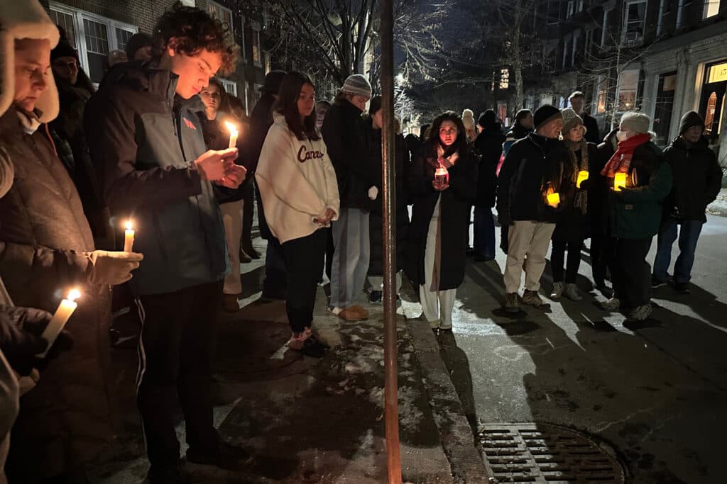 A group of people stand outside at night holding lit candles during a vigil. They are dressed in winter clothing, suggesting a cold night. The street is dimly lit, with the warm glow of the candles illuminating their faces.