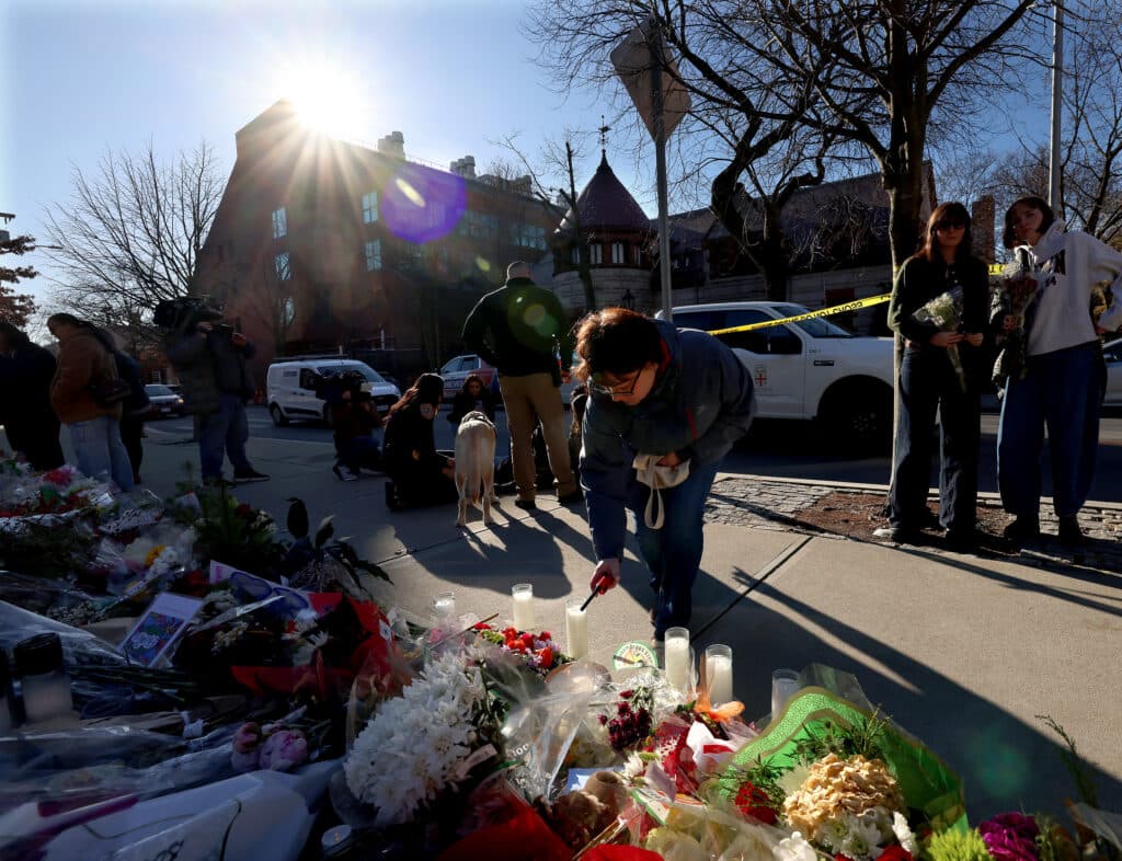 People gather at a memorial site with flowers and candles on the sidewalk, under bright sunlight, with a film crew and police tape visible in the background.