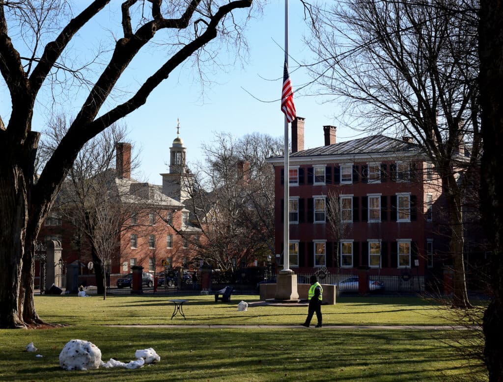 A person wearing a bright jacket walks across a grassy area in front of historical brick buildings, with an American flag flying at half-mast on a clear day, surrounded by leafless trees and patches of snow on the ground.
