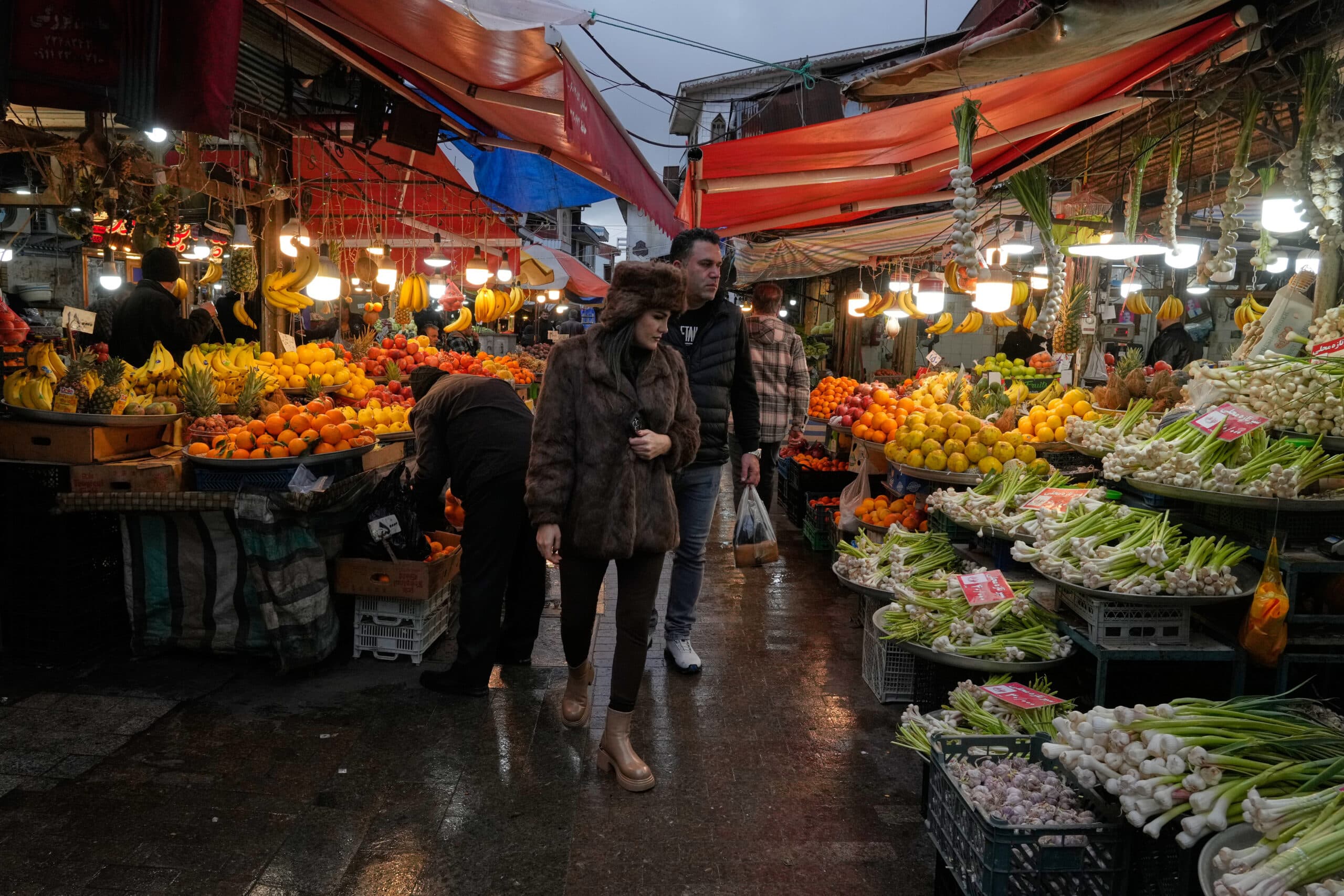 People walking through a busy market filled with vibrant displays of fruits and vegetables, including bananas, oranges, and green onions, under colorful awnings.