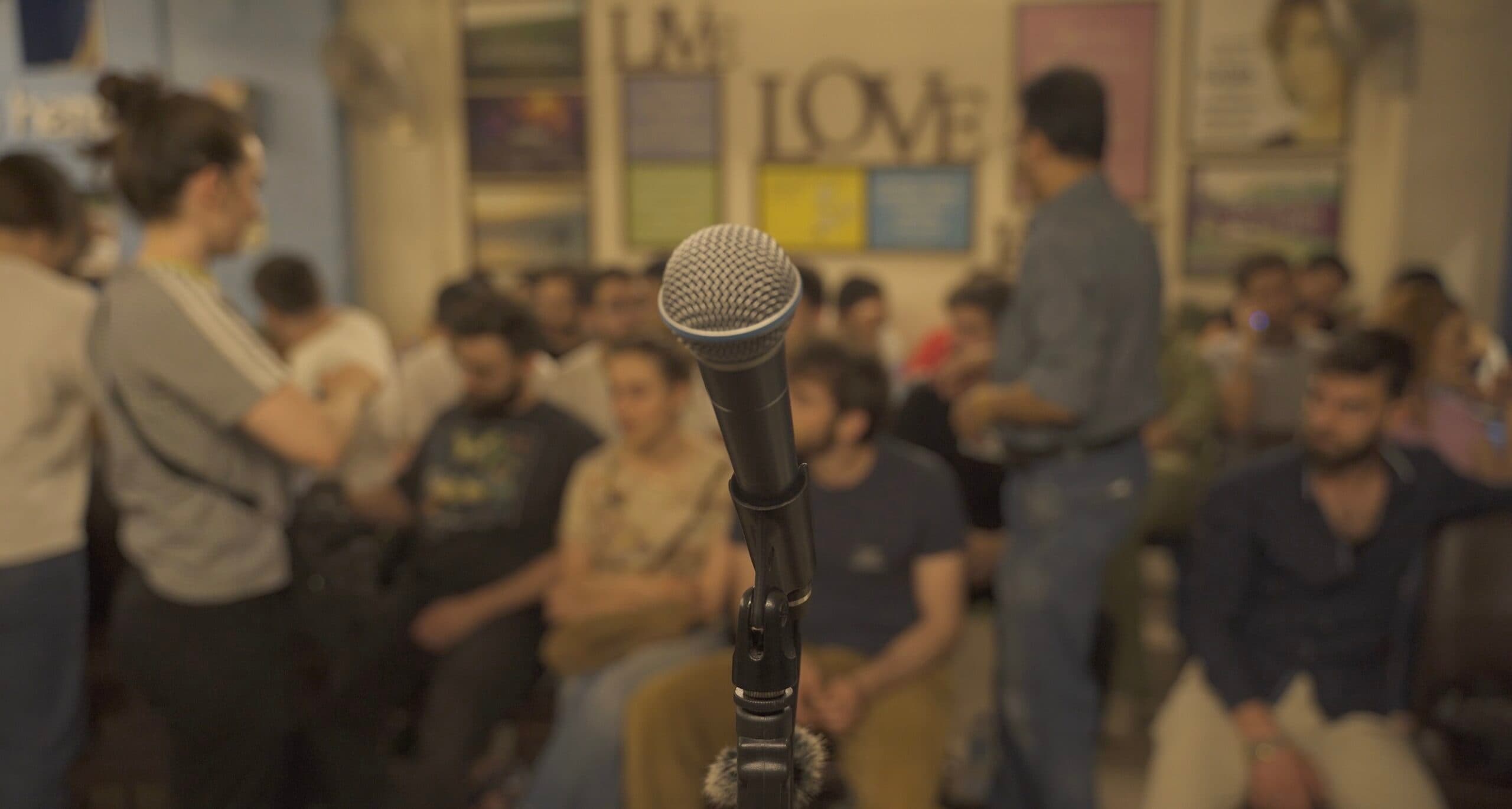 A close-up of a microphone with a blurred background showing a seated audience in a casual indoor setting, with colorful posters on the walls and people engaging in conversation.