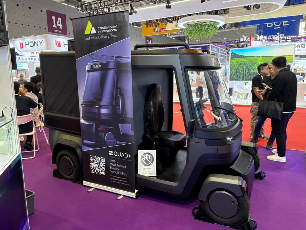 A futuristic smart modular electric vehicle displayed on a purple carpet at a trade show, with promotional signage by Castle Peak Innovations. Several people are standing nearby, and the background shows other exhibits and booths.