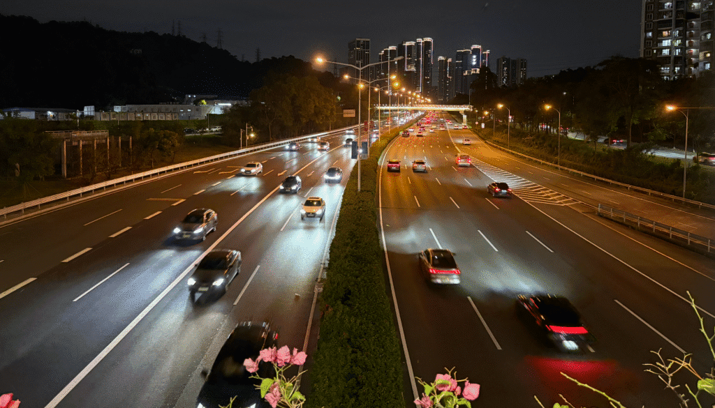 A nighttime view of a busy highway with cars moving in both directions, surrounded by city buildings with lights illuminating the scene.