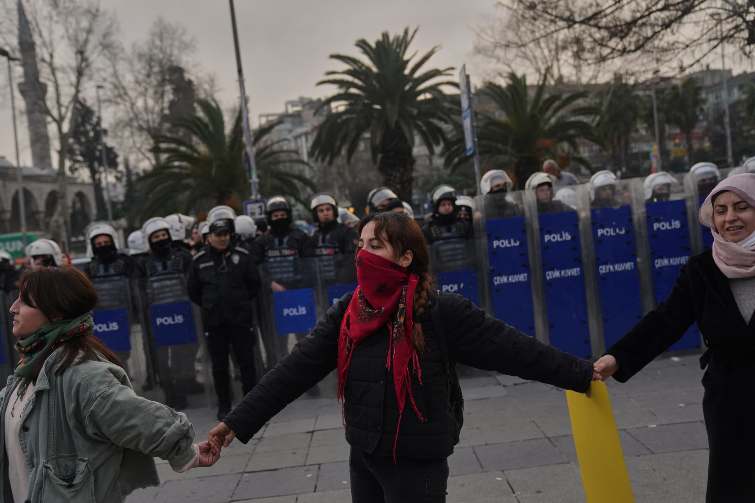 Protesters holding hands form a human chain in front of a line of riot police wearing helmets and standing behind shields, in an outdoor setting with palm trees in the background.