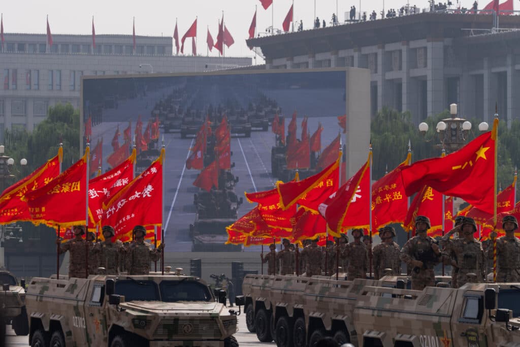 Military parade with soldiers standing on armored vehicles, holding red flags with yellow text, in front of a screen displaying tanks and additional flags.
