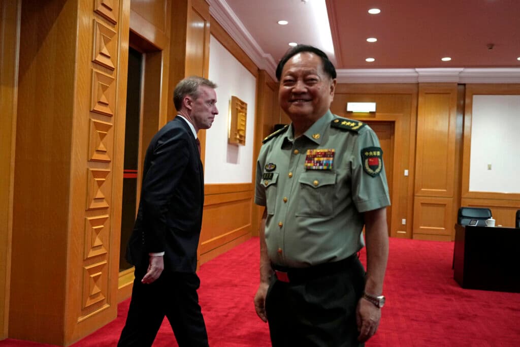 A smiling man in a military uniform stands in a room with wooden paneling, while another man in a suit walks past him.