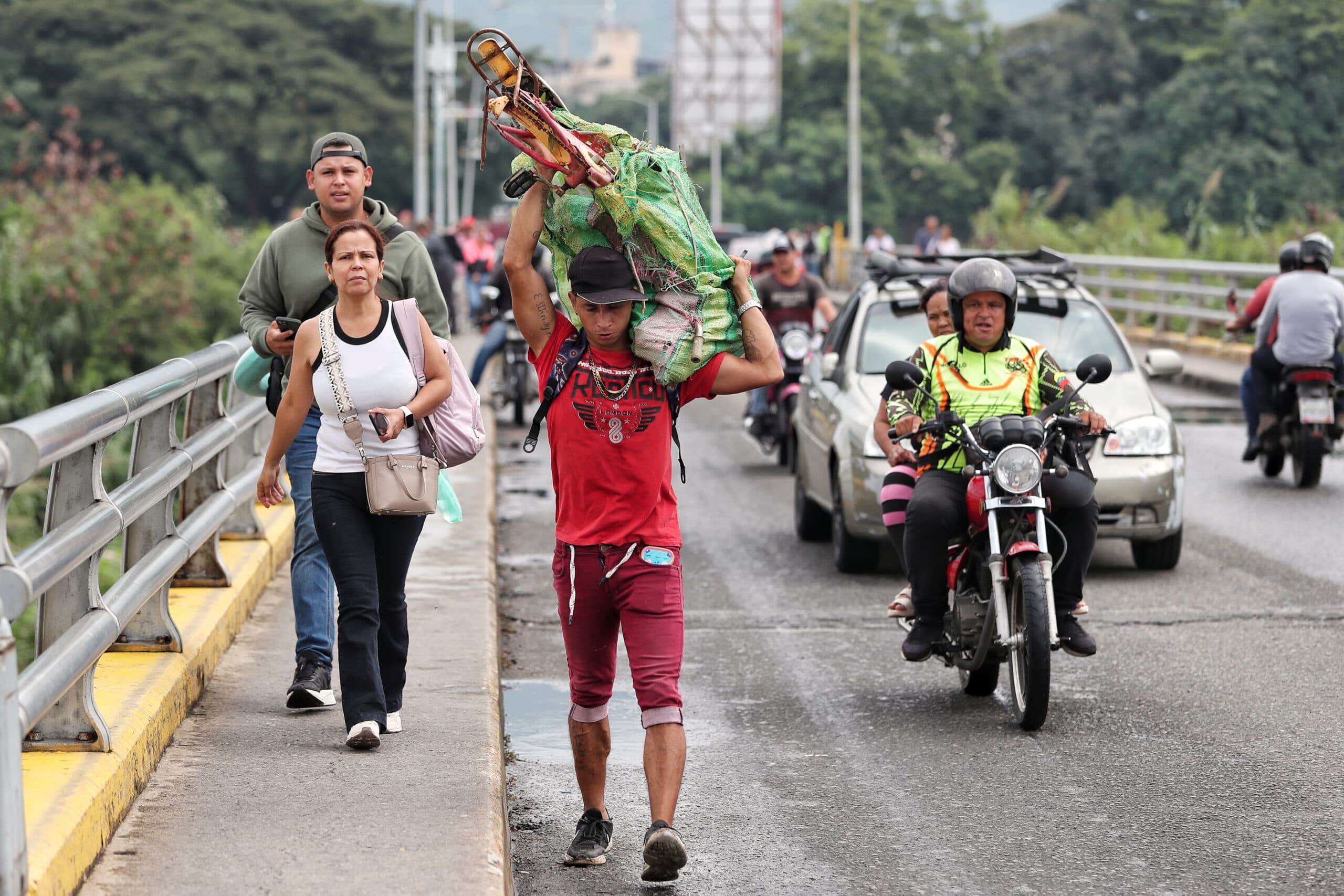 A man in a red shirt carries a large bundle on his shoulders while walking on a bridge. He is accompanied by others on foot, and motorcycles and cars drive by on the road next to them. The setting appears to be an urban area with trees in the background.