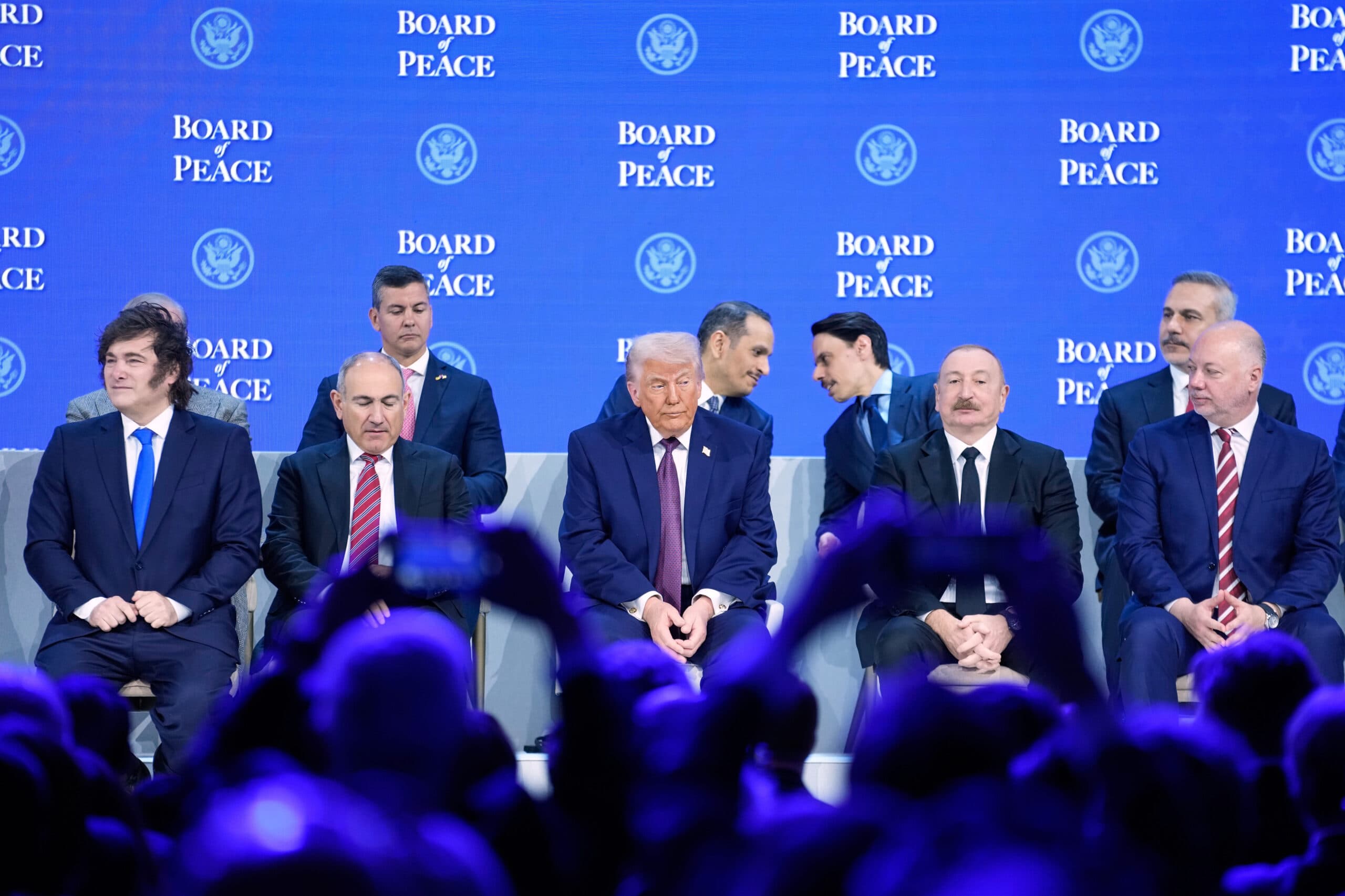 A group of men in suits sit on a stage in front of a blue backdrop with the text "Board of Peace" repeated. The scene is framed by an audience, some holding up phones.